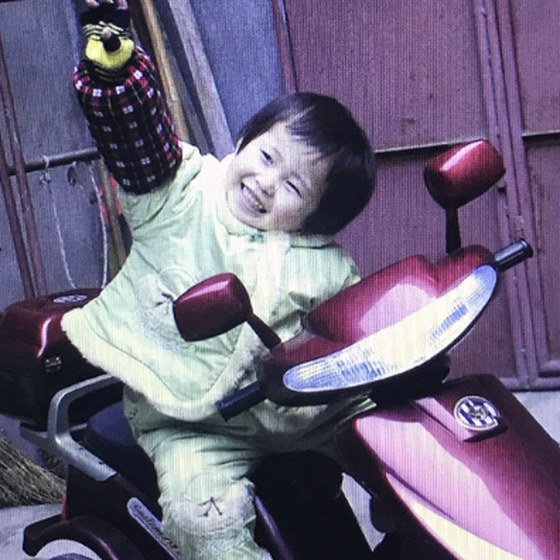 Connie as a toddler, grinning on a toy motorbike with an arm raised